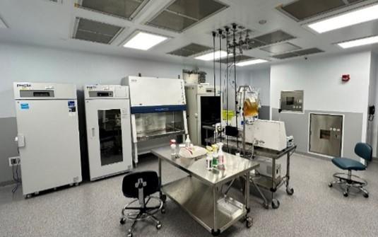 A laboratory room with various scientific equipment. There are large white laboratory machines lined up against the walls, including incubators and a biosafety cabinet. In the center, a stainless steel worktable is equipped with lab supplies such as bottles and containers. There are two chairs with wheels near the table. The room has overhead lights and the floor is covered with speckled gray tiles. 