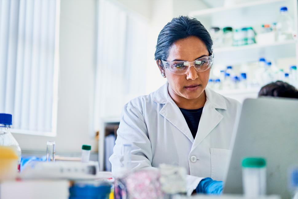 A researcher in a lab coat and goggles working on a laptop in a lab.