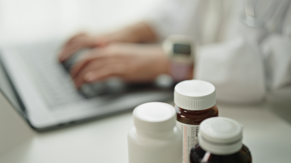 A doctor wearing a white coat and stethoscope types on a laptop near three closed medicine bottles.