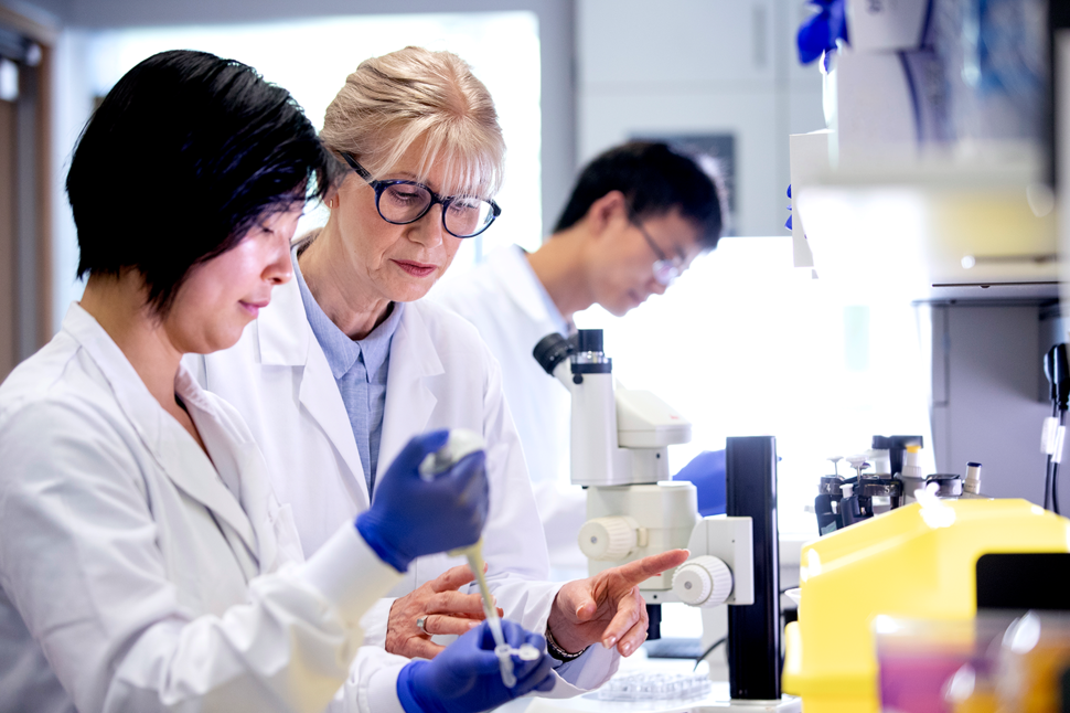 Three researchers - two women and one man - in a lab environment. The two women in the foreground wear white lab coats; one observes another putting a sample into a tube. The man is blurry in the background. A microscope and other lab equipment are near the women. 