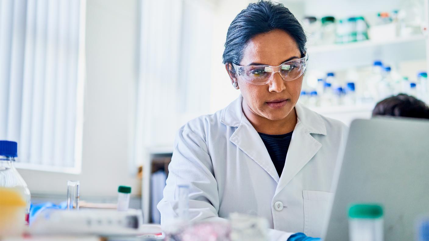 A researcher in a lab coat and goggles working on a laptop in a lab.