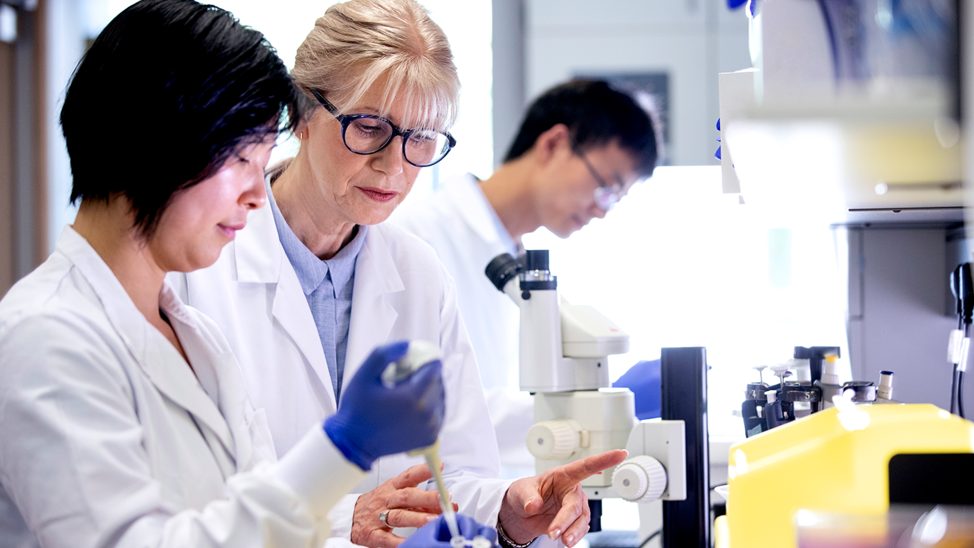 Three researchers - two women and one man - in a lab environment. The two women in the foreground wear white lab coats; one observes another putting a sample into a tube. The man is blurry in the background. A microscope and other lab equipment are near the women. 