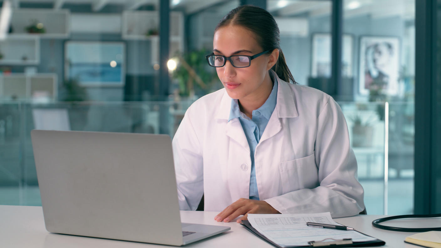 A woman in a white lab coat is sitting at a desk in front of a laptop in an office.