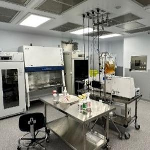 A laboratory room with various scientific equipment. There are large white laboratory machines lined up against the walls, including incubators and a biosafety cabinet. In the center, a stainless steel worktable is equipped with lab supplies such as bottles and containers. There are two chairs with wheels near the table. The room has overhead lights and the floor is covered with speckled gray tiles. 